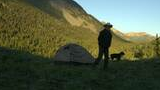 Person wearing a cowboy hat standing near a small tent in a grassy clearing with a black and white dog, surrounded by large mountains covered in dense evergreen trees under a clear sky