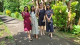 Four young women standing together on a shaded paved path surrounded by green foliage and banana plants, two of them showing peace signs with their hands