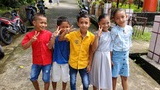Five children standing on a paved path outdoors, smiling and making peace signs, with scooters and greenery in the background