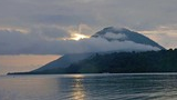 Sun partially hidden behind clouds over a forested mountain with a calm body of water reflecting the light in the foreground