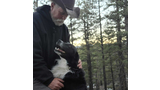 Older man in a cowboy hat and dark hoodie kneeling and looking affectionately at a black and white dog sitting close and looking up at him in a forested area with pine trees and patches of snow