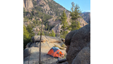 Orange and gray tent pitched on a rocky and grassy campsite surrounded by large boulders and pine trees with a mountain covered in rocks and trees in the background under a blue sky