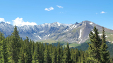 Snow-capped mountain range with green pine trees in the foreground under a blue sky with scattered clouds