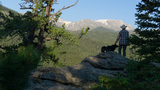 Person wearing a hat and plaid shirt standing on a large rock with a black dog, overlooking a forested valley and snow-capped mountains in the distance