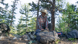 Man in a plaid shirt and cowboy hat kneeling on the ground in a forest clearing, packing or unpacking a large backpack, with three dogs nearby resting on the pine needle-covered forest floor