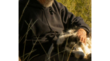 Man wearing dark clothing and a wide-brimmed hat gently stroking a black and white dog resting its head on his lap amid tall grass and logs