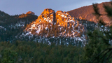 Rocky mountain peak partially covered with snow, glowing orange in the light of the setting or rising sun, surrounded by dense forest