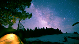 a glowing tent set up in a dark wooded area with snow patches, beneath a night sky filled with stars and the bright arc of the Milky Way