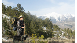Man standing on a mountain trail with a hiking backpack, accompanied by a black dog wearing a harness, with snow-capped peaks and dense trees in the background