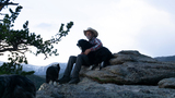 Man sitting on a large rock outdoors hugging a black and white dog with another dog walking nearby and mountains in the background