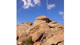 sunlit large weathered granite rock formations with some small trees and shrubs against a blue sky with scattered clouds
