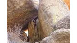 Narrow passage formed by large, weathered boulders and rock formations with some dry shrub branches at the base