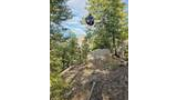A black backpack hanging suspended in the air between trees in a forested area with a large rock and scattered branches on the ground
