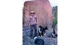 Man wearing a cowboy hat and plaid shirt standing in front of a large pinkish rock formation with two black and white dogs nearby in a natural outdoor setting