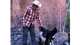 Man wearing a cowboy hat and plaid shirt petting a sitting black and white dog in a natural outdoor setting with leafless trees and a reddish rock wall background