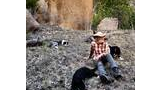 man wearing a cowboy hat sitting on a grassy slope with three dogs resting nearby and large rock formations in the background