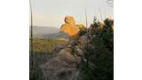 Sunlit rock formation resembling a profile of a face overlooking a forested valley with distant mountains and a pale sky