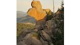 Large rock formation bathed in warm sunlight, shaped like a sitting bear, surrounded by pine trees and distant mountain ranges under a pale sky