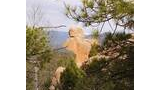 Large tan rock formation resembling a balanced figure surrounded by green pine and deciduous trees with distant snow-capped mountains under a partly cloudy sky