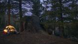 Illuminated tent pitched among tall pine trees and large rocks at dusk in a forested mountain area