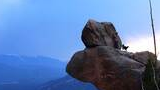 Person wearing a cowboy hat standing with a black and white dog atop a large rock formation overlooking a mountain landscape under a blue sky