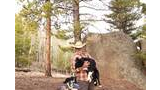 Man in a cowboy hat holding a black and white dog in a forested area with pine trees and large rocks, with two other dogs resting on the ground nearby