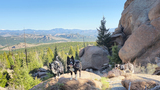 Two border collie dogs wearing backpacks standing on rocky terrain with a third dog resting near large boulders, overlooking a forested mountainous landscape under a clear blue sky