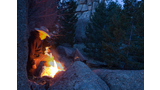 Person in a cowboy hat tending a campfire among large rocks and pine trees at dusk