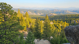 View of a forested mountain landscape with large boulders in the foreground and distant snow-capped peaks under a clear sky