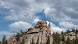 Large rugged rock formation surrounded by green pine trees under a partly cloudy sky