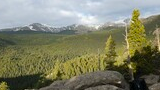 Rocky outcrop overlooking a vast forested valley with snow-capped mountains in the distance and a black and white dog standing among the rocks