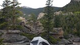 A tent pitched on rocky ground surrounded by pine trees and large boulders with a mountainous forest landscape in the background, and a black and white dog resting nearby