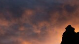 Silhouetted rocky ridge and tree-lined horizon against a dramatic sunset sky with dark clouds and warm orange light