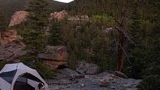 Two dogs near a tent set up on rocky terrain surrounded by pine trees with forested mountains and a rocky peak in the background at dusk