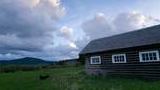 Historic wooden cabin with three white-trimmed windows in a grassy field under a sky filled with large clouds during early evening