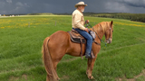Man wearing a cowboy hat and beige jacket riding a chestnut horse on a grassy field under a dark, cloudy sky