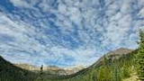 Mountain basin with dense evergreen forest, rocky peaks with patches of snow, under a sky filled with scattered white clouds on a clear day