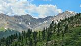Mountain landscape with green forested slopes, rocky peaks with patches of snow, and a partly cloudy blue sky