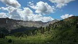 lush green alpine valley with scattered evergreen trees under a partly cloudy blue sky, surrounded by rocky mountain peaks with patches of snow