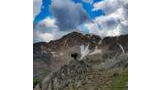 Black dog standing on rocky outcrop with a mountainous ridge behind and partly cloudy sky overhead