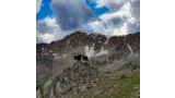 Two black dogs standing on a rocky outcrop with rugged mountain peaks and patches of snow under a partly cloudy blue sky