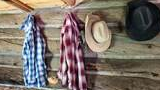Two plaid shirts and two cowboy hats hanging on the interior wooden wall of a rustic cabin