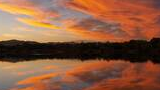 Vibrant orange and purple clouds reflecting in a calm lake at sunset with a distant mountain range and tree-lined shore