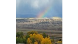 Landscape with autumn trees in the foreground, an expansive valley with scattered trees and bushes, and a rainbow appearing against a cloudy sky above distant hills