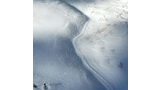 Snow-covered ground with smooth, glistening curves and shadows highlighting the texture, small dried plants emerging through the snow