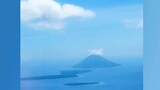 Island surrounded by calm blue water with a lone mountain rising in the distance under a blue sky with scattered clouds
