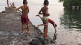 Children standing and playing along a rocky pier next to calm water with trees on the right side and a hazy background