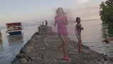 children standing and walking on a rocky pier extending into calm water with boats docked nearby under a partly cloudy sky at sunset
