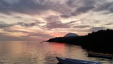 Calm water at sunset with two small boats anchored near a forested shoreline and a mountain silhouette in the distance under a cloud-filled sky