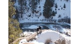 Black dog standing on a snow-covered rock overlooking a partially frozen river surrounded by snow and pine trees